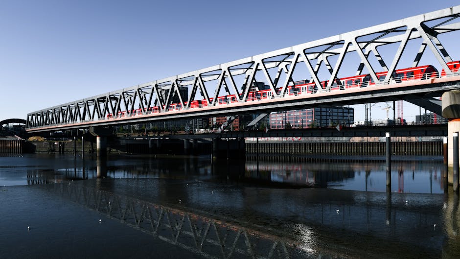 A modern railway bridge with a white steel framework and red train cars crossing over a calm body of water in an urban setting. The bridge features a truss design with triangular sections, supported b