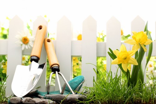 A close-up view of a small outdoor garden area featuring a white picket fence as the background. In the foreground, there are two gardening tools—a trowel and a hand fork—both with wooden handles and metal working ends—resting inside a pair of gardening gloves. To the right, vibrant yellow daffodils and white daisies grow amidst lush green grass and foliage. The garden surface consists of dark soil and small patches of grass, with some pebbles scattered nearby. The scene is illuminated by natural daylight, highlighting the bright colours of the flowers and the crisp white of the fence, creating a welcoming and well-maintained outdoor space typical of a residential garden that could be serviced by Gardener Deptford in the nearby area postcode. The overall environment appears fresh and well-kept, ideal for planting, maintenance, or garden tidying activities, reflecting professional gardening and landscaping practices.